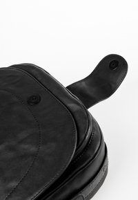 Mustavärinen nahkatasku, jossa on sileä pinta, kaareva läppämuotoilu ja nappisuljin. Pohjassa on hillitty kohokuviollinen yksityiskohta.