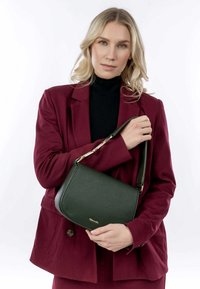 Grön texturerad läderhandväska med ett guldaccent och justerbar rem, hålls av en person som bär en bordeauxfärgad kavaj och svart tröja.