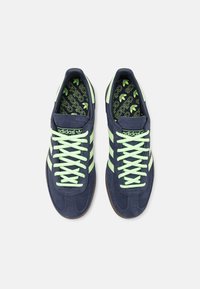 Marinblå mockaskor med ljusgröna accenter, som har tre ränder, runda snören och texturerade gummisulor. Interiören har logomönster.