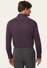 Langärmliges Hemd aus dunkelviolettem Stoff, mit klassischem Kragen und Knopfmanschetten, kombiniert mit grauen Hosen. Glatte Textur.