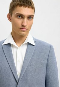 Blazer texturé bleu clair avec un col cranté, une poche poitrine unique, porté par-dessus une chemise blanche à boutons. Gros plan sur le haut du corps.