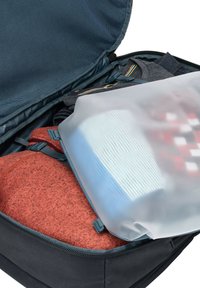 Zwarte rugzak interieur met opgevouwen rode en grijze kleding, gedeeltelijk bedekt door een doorzichtige zak die een blauw item met geribbelde textuur vasthoudt.