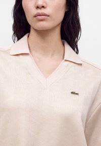 Femme portant un polo texturé rose pâle avec un petit logo crocodile vert sur la poitrine gauche, montré des épaules jusqu'aux lèvres.