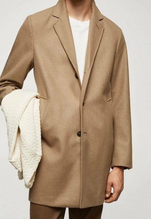 Uomo che indossa un cappotto color cammello con bottoni e una camicia bianca, con un maglione lavorato a trecce color crema piegato sul braccio.