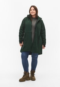 Donkergroene hooded jacket met een rits aan de voorkant, zijzakken en zwarte afwerking. Gecombineerd met blauwe jeans en bruine veterschoenen.