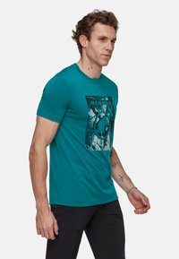 Turkusowy t-shirt z krótkim rękawem wykonany z bawełny, z niebiesko-białym nadrukiem graficznym oraz napisem "MAMMUT" na środku. Gładka faktura.
