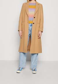 Długi beżowy płaszcz z dwoma przednimi kieszeniami i klapami, nałożony na wielokolorowy sweter z dzianiny oraz jasnoniebieskie spodnie typu wide-leg.