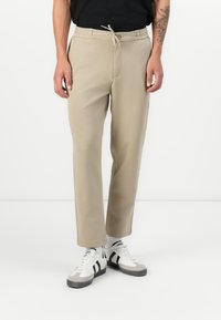 Beige-väriset collegehousut med elastisk midja, snörning, sidofickor och avslappnad passform, tillsammans med vita sneakers med svarta detaljer.