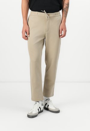 Beige sweatpants with an elastic waistband, drawstring, side pockets, and a relaxed fit, paired with white sneakers featuring black accents.