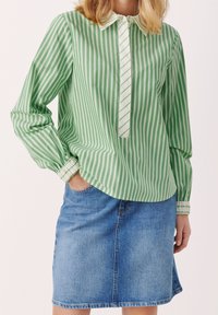 Zeleno-bílý pruhovaný košile s klasickým límcem a knoflíkovou légu, spárovaná s modrou denimovou sukní v tužce. Dlouhé rukávy s manžetami.