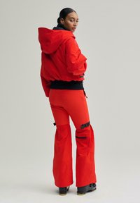 Geacă roșie cu glugă, căptușită și manșete elastice, și pantaloni asortați mulați. Pantalonii au accente negre și picioare largi evazate. Cizme negre vizibile.