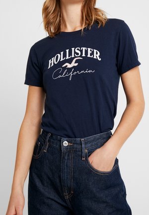 Kobieta w granatowym T-shircie Hollister California schowanym w ciemnoniebieskie jeansy, stojąca z jedną ręką w kieszeni na jasnym tle.
