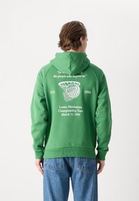 Πράσινη hooded μπλούζα με λευκή εκτύπωση γραφικών στην πλάτη που περιλαμβάνει κείμενο και σχέδιο ενός μπαστουνιού λακρός. Ριχτές μανσέτες και στρίφωμα.