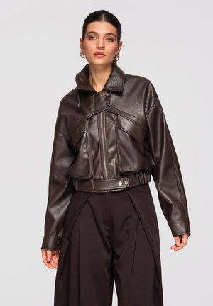 Giovane donna che indossa una giacca di pelle marrone scuro con texture e collo alto, abbinata a pantaloni neri a gamba larga, su uno sfondo chiaro e uniforme.