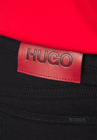 Los jeans negros presentan una etiqueta de cuero roja con "HUGO" en relieve. Se puede apreciar una textura suave, un corte recto y detalles de costura mínimos.