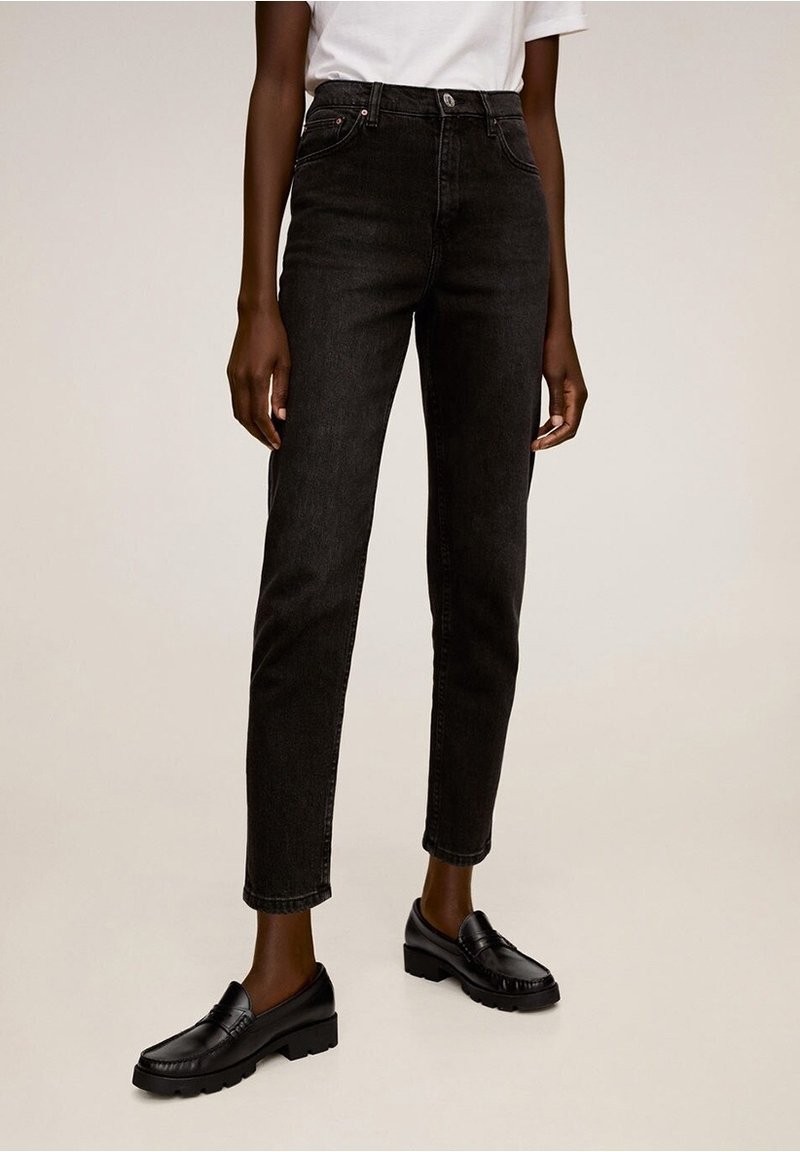 Personne portant un jean noir taille haute, une chemise blanche rentrée, et des mocassins noirs, debout sur un fond clair uni.