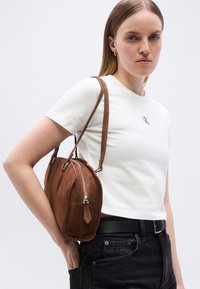 Γυναίκα που φοράει λευκό κοντό μπλουζάκι και μαύρο τζιν, κρατώντας καφέ σουέντ τσάντα στον ώμο, μπροστά σε απλό φόντο.