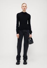 Crna rebrasta turtleneck majica uparenjena s tamnim farmerkama i debelim crnim cipelama. U jednoj ruci drži malu crnu torbicu s umetkom od rupica.