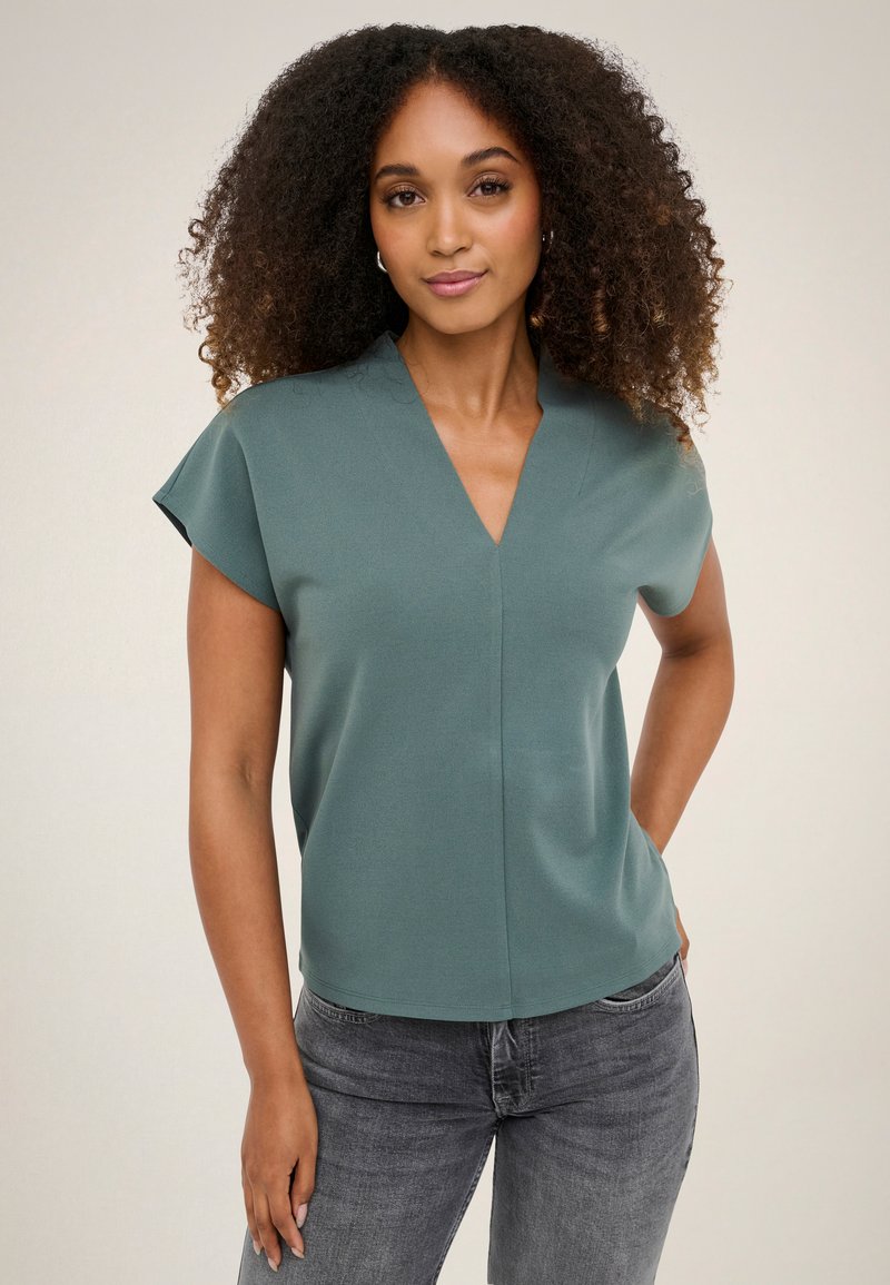 Femme aux cheveux bouclés portant un top vert à manches courtes et col en V ainsi qu’un jean gris, debout, une main dans la poche, sur un fond uni.
