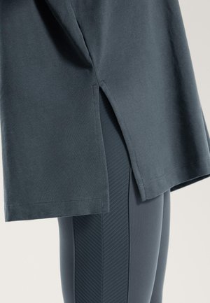 Sötét szürke pamut póló oldalvágásokkal, sötét szürke leggingssel párosítva, amelyek texturált függőleges csíkmintázattal rendelkeznek.