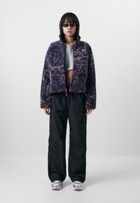 Giacca in pile viola e nera con motivo, con zip frontale, abbinata a pantaloni neri larghi e sneakers argento. Dettaglio con coulisse rosa regolabile.