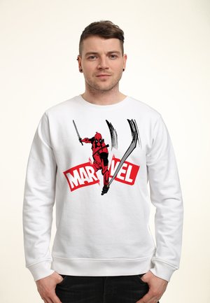 Mężczyzna w białej bluzie z czerwoną i czarną postacią Deadpoola oraz logo Marvel, stoi z rękami w kieszeniach na jednolitym tle.