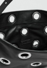 Interior de una bolsa de cuero negro que presenta ojales plateados. Textura suave con costuras visibles y una pequeña etiqueta de marca. Bordes lisos y redondeados.