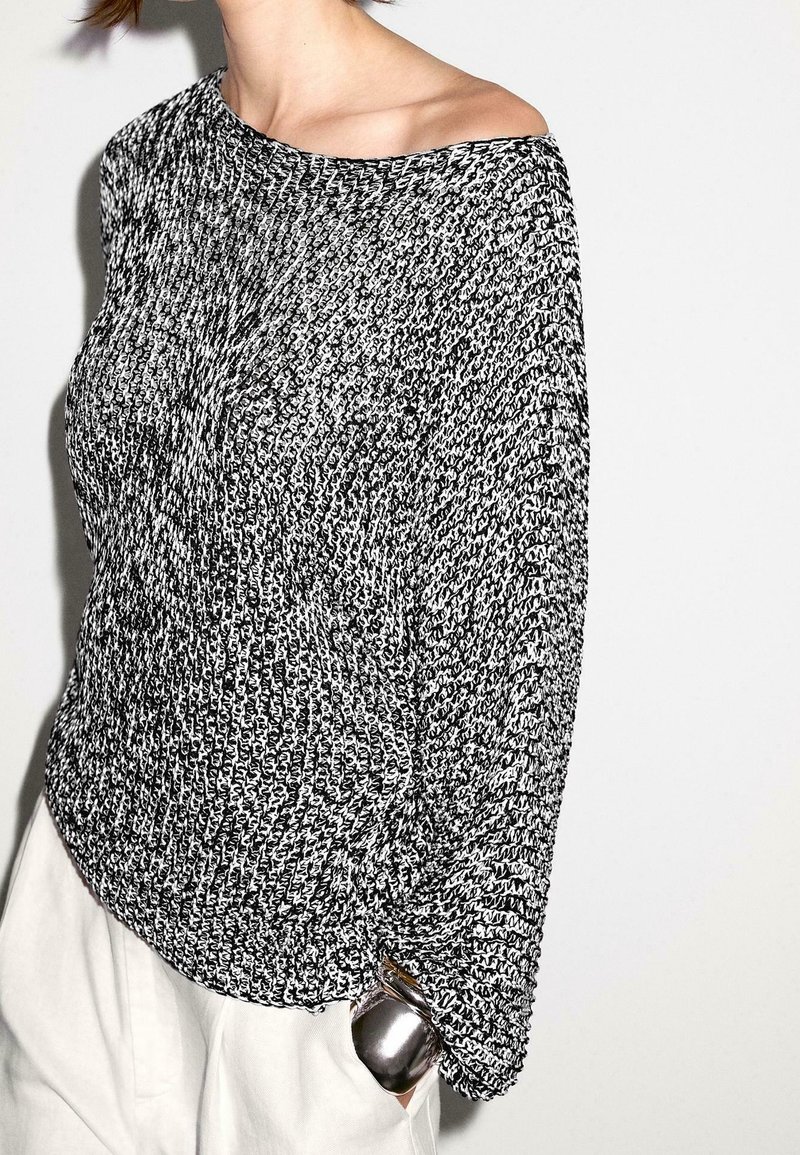 Black and white knitted sweater with an asymmetrical neckline, loose fit, and textured fabric. Silver hardware detail on the side.