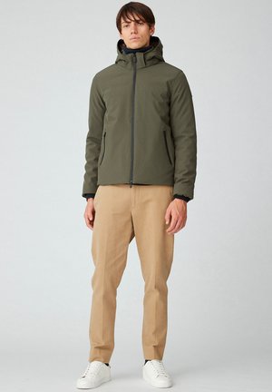 Giacca a vento verde oliva con cappuccio, zip frontale, tasche laterali e dettagli neri, abbinata a pantaloni beige e sneakers bianche.