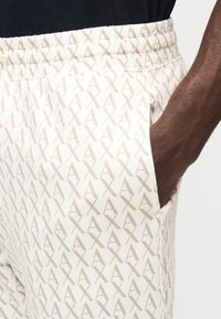 Beżowe joggery w wzory z elastyczną talią i bocznymi kieszeniami, z subtelnym powtarzającym się motywem logo w jaśniejszym odcieniu.