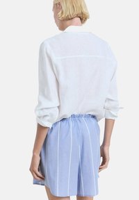 Chemise en lin blanche à manches retroussées, associée à un short bleu clair avec des rayures verticales blanches et une taille élastique.