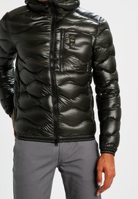 Schwarze, wattierte Jacke mit Reißverschluss vorne, strukturiertem Wellenmuster und einer Brusttasche mit kleinem Logo. Glatte, glänzende Oberfläche.