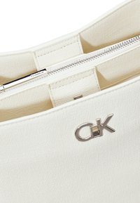 Borsa a tracolla bianca con texture, dotata di hardware argentato, chiusura a zip e un'accents con il logo "CK" sulla parte anteriore. L'interno presenta più scomparti.