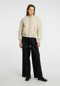 Una donna sta in piedi con sicurezza, indossando un bomber beige chiaro e pantaloni neri larghi, con capelli lunghi e lisci e un'espressione seria.