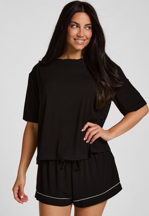 Vrouw met lang donker haar, gekleed in een losse zwarte T-shirt en bijpassende shorts met witte bies, staand met hand op heup en glimlachend, kijkend naar de zijkant.