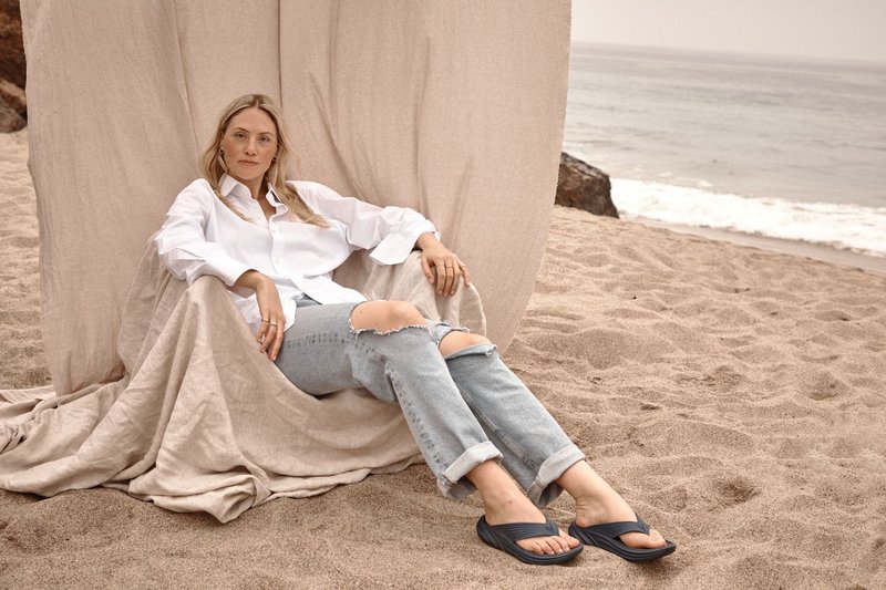Femme en chemise blanche et jean déchiré se détendant sur un fauteuil en tissu beige sur une plage de sable près des vagues de l'océan.