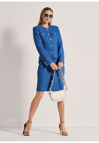 Blauwe textuurblazer met gouden knopen, bijpassende rok en witte handtas. Model draagt zonnebril en zwarte hakken met bandjes.