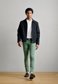 Μαύρο μπουφάν bomber πάνω από λευκό πόλο πουκάμισο, συνδυασμένο με πράσινο chino και σκούρα loafers. Ομαλές υφές και εφαρμογή προσαρμοσμένη σε όλο το σύνολο.