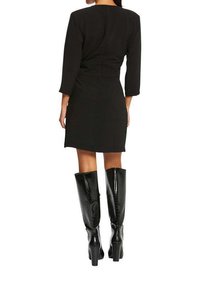 Robe noire à manches trois quarts, design fuselé et fermeture éclair dans le dos. Associée à des bottes noires en cuir verni montantes avec des talons.