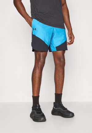 Homme portant un short de sport noir et bleu avec des baskets et des chaussettes noires, debout devant un fond clair uni.