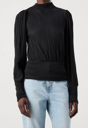 Persoon draagt een zwart geplooid shirt met lange mouwen en een hoge hals, gecombineerd met lichtblauwe spijkerbroek.