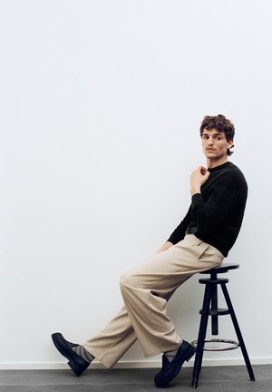 Giovane uomo con capelli ricci che indossa pantaloni beige, maglione nero e scarpe nere, seduto di lato su uno sgabello nero regolabile contro una parete bianca.