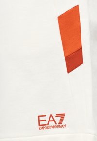 Λευκό ύφασμα με κόκκινες γραμμές σε γωνία και κεντημένο λογότυπο EA7 Emporio Armani κοντά στην κάτω άκρη.