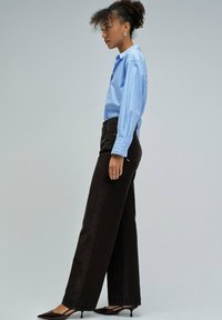 Camisa de botones azul claro, pantalones anchos marrones de tela texturizada y tacones negros de punta. El modelo está de perfil contra un fondo gris.