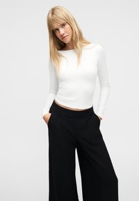 Νεαρή γυναίκα με ξανθά μαλλιά στέκεται με τα χέρια στις τσέπες, φορώντας λευκή μπλούζα με μακριά μανίκια και μαύρο παντελόνι με φαρδιά μπατζάκια σε απλό φόντο.