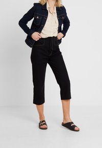 Veste en jean bleu foncé avec boutons en laiton, chemise beige à boutons, pantalons noirs courts avec couture contrastante et sandales noires à enfiler.