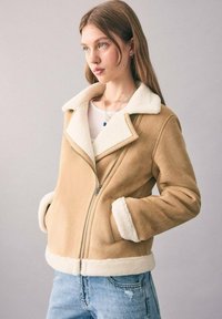 Beige Faux-Suede-Jacke mit einem weißen Lammfellkragen und -manschetten, ausgestattet mit einem diagonalen Reißverschluss und Seitentaschen. Getragen über einem weißen Oberteil.