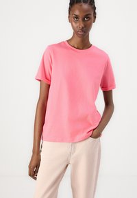 Roze katoenen T-shirt met korte mouwen, een ronde halslijn en een ontspannen pasvorm, gecombineerd met lichtgekleurde broek. Soepele textuur, minimalistisch ontwerp.