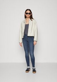 Chaqueta blanca corta, camiseta de tirantes gris, pantalones vaqueros ajustados azules, zapatillas negras con suela de goma y gafas de sol rectangulares negras. Fondo plano.