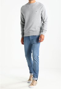 Sudadera gris de cuello redondo con puños y dobladillo acanalados, llevada con jeans azul claro y zapatillas beige con suelas blancas. Detalle de logo sencillo.